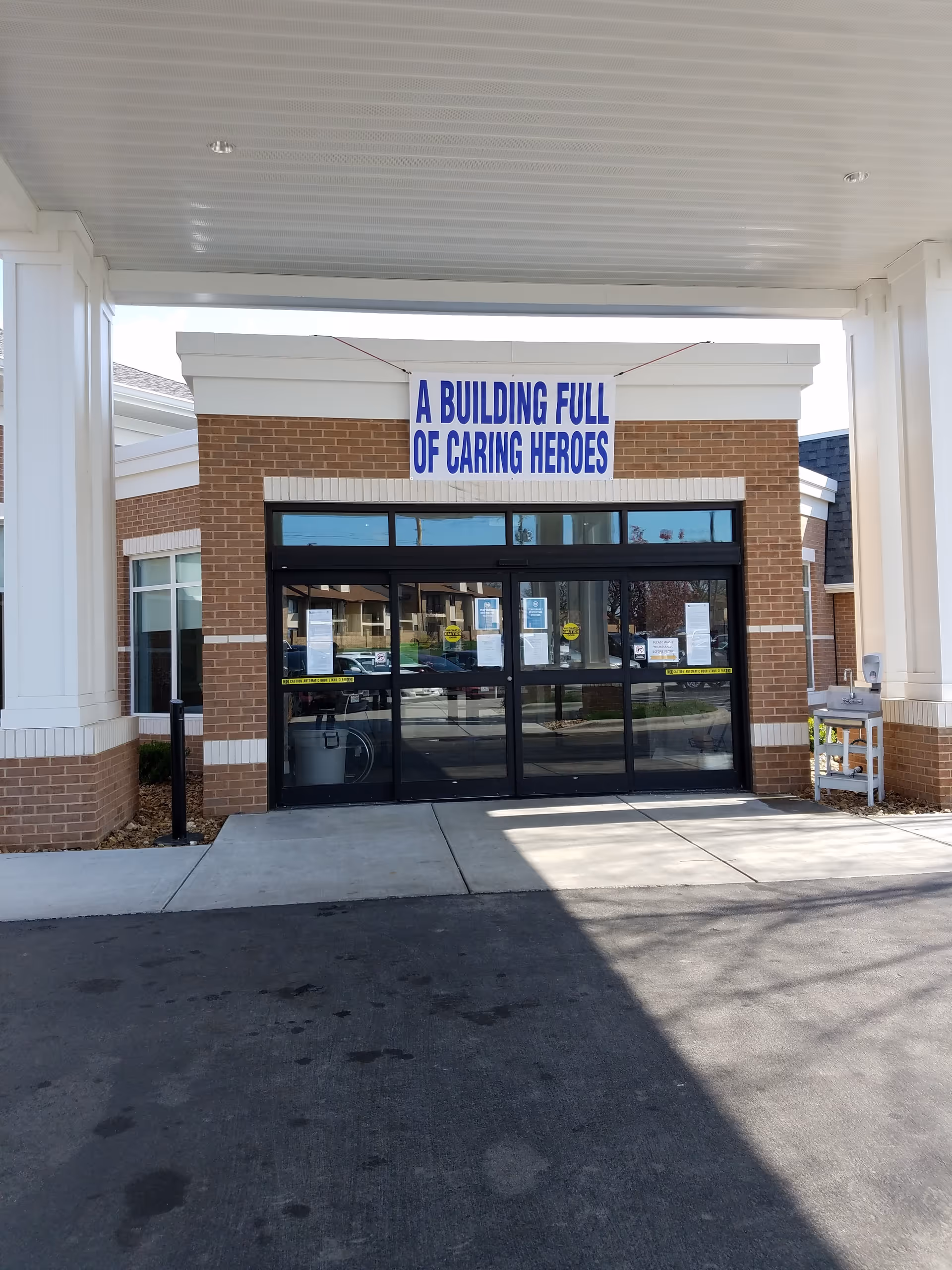 Entrance of a brick building with glass automatic sliding doors. Above the doors is a large white sign with blue text that reads 'A BUILDING FULL OF CARING HEROES'. The entrance is covered by a white canopy supported by columns.