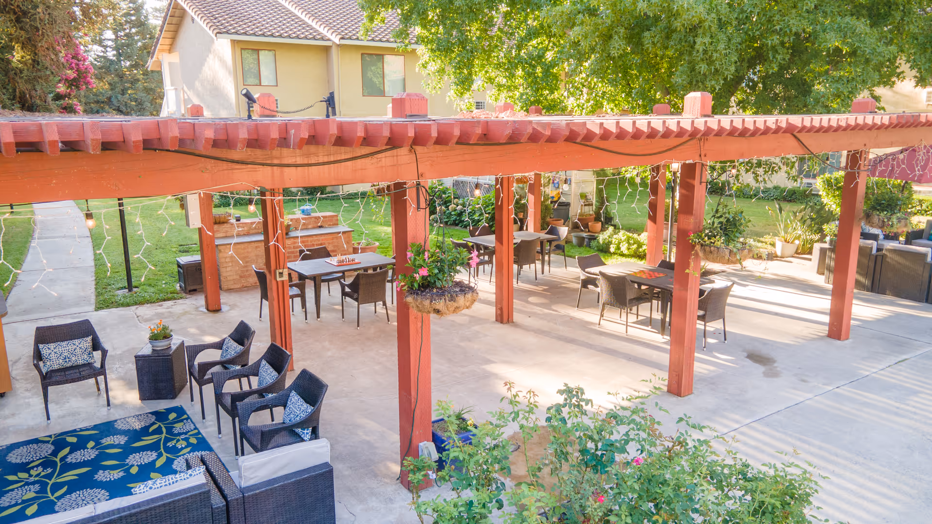 Covered outdoor patio with a wooden pergola, tables, chairs, and potted plants in a landscaped courtyard.