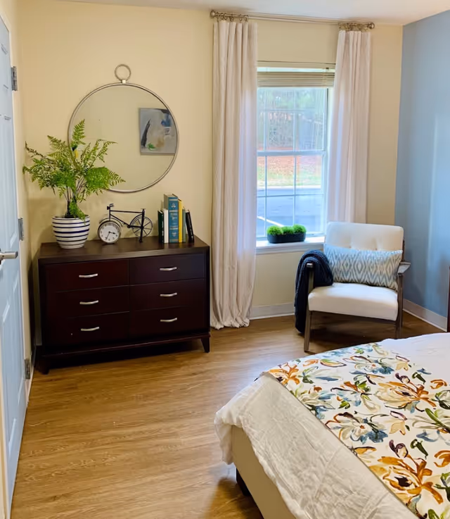 A cozy bedroom with a wooden dresser topped with a potted plant, a decorative bicycle, a clock, and books. Above the dresser is a round mirror and a small abstract painting. Next to the dresser is a window with white curtains, a small green plant on the windowsill, and a white cushioned chair with a patterned pillow and a dark throw draped over one arm. Part of a bed with a colorful floral bedspread is visible in the foreground. The room has light-colored walls and wood flooring.