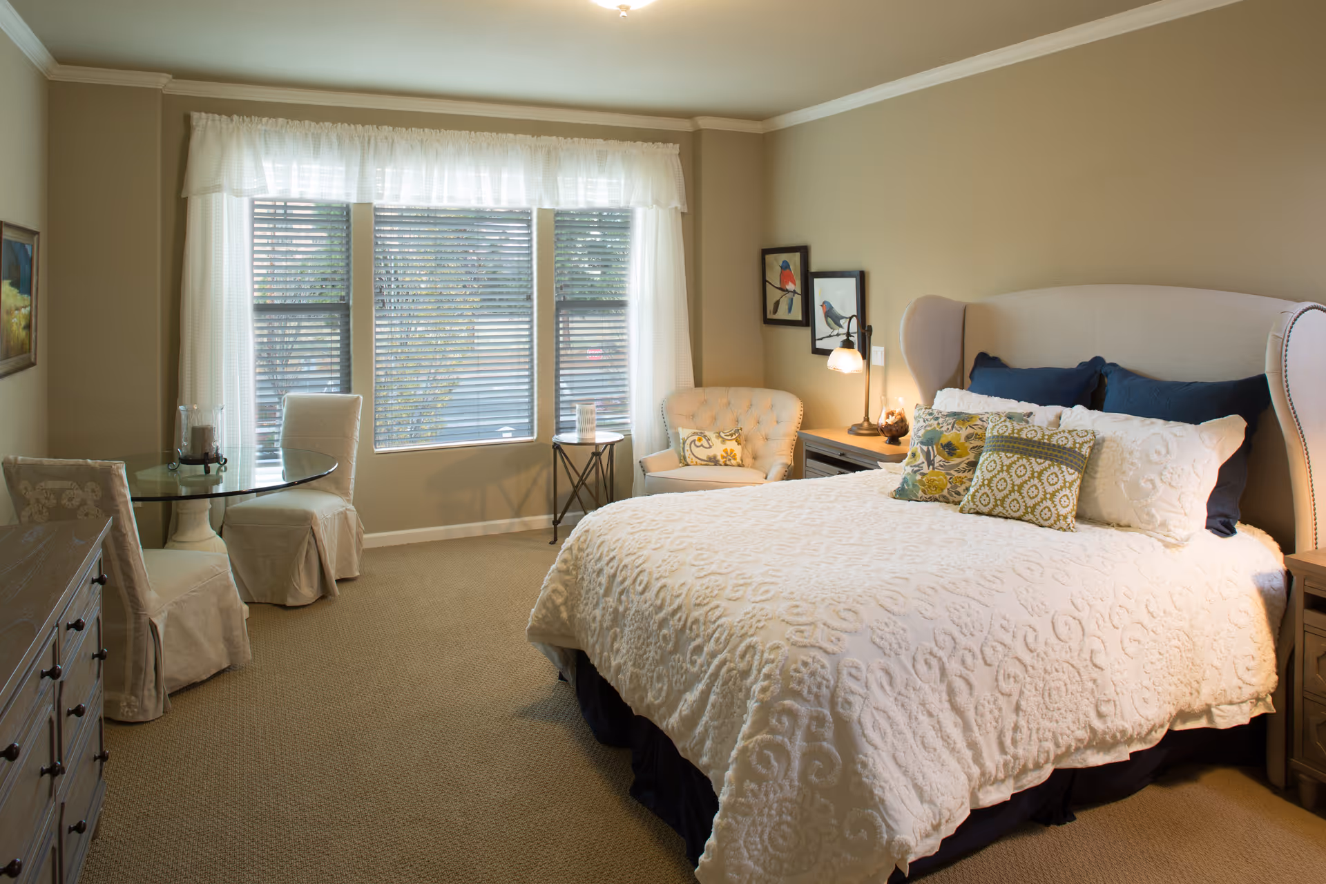 Well-lit bedroom with a large bed, tufted headboard, nightstands, and seating near a window.