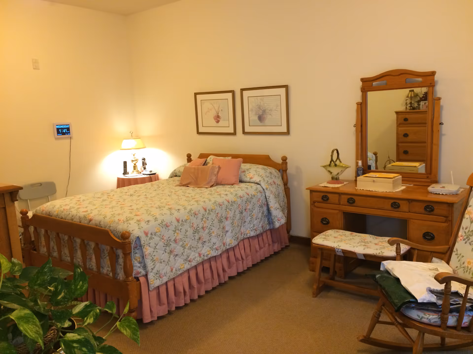 A cozy bedroom with a wooden bed dressed in floral bedding, matching wooden dresser with mirror, nightstand and seating.