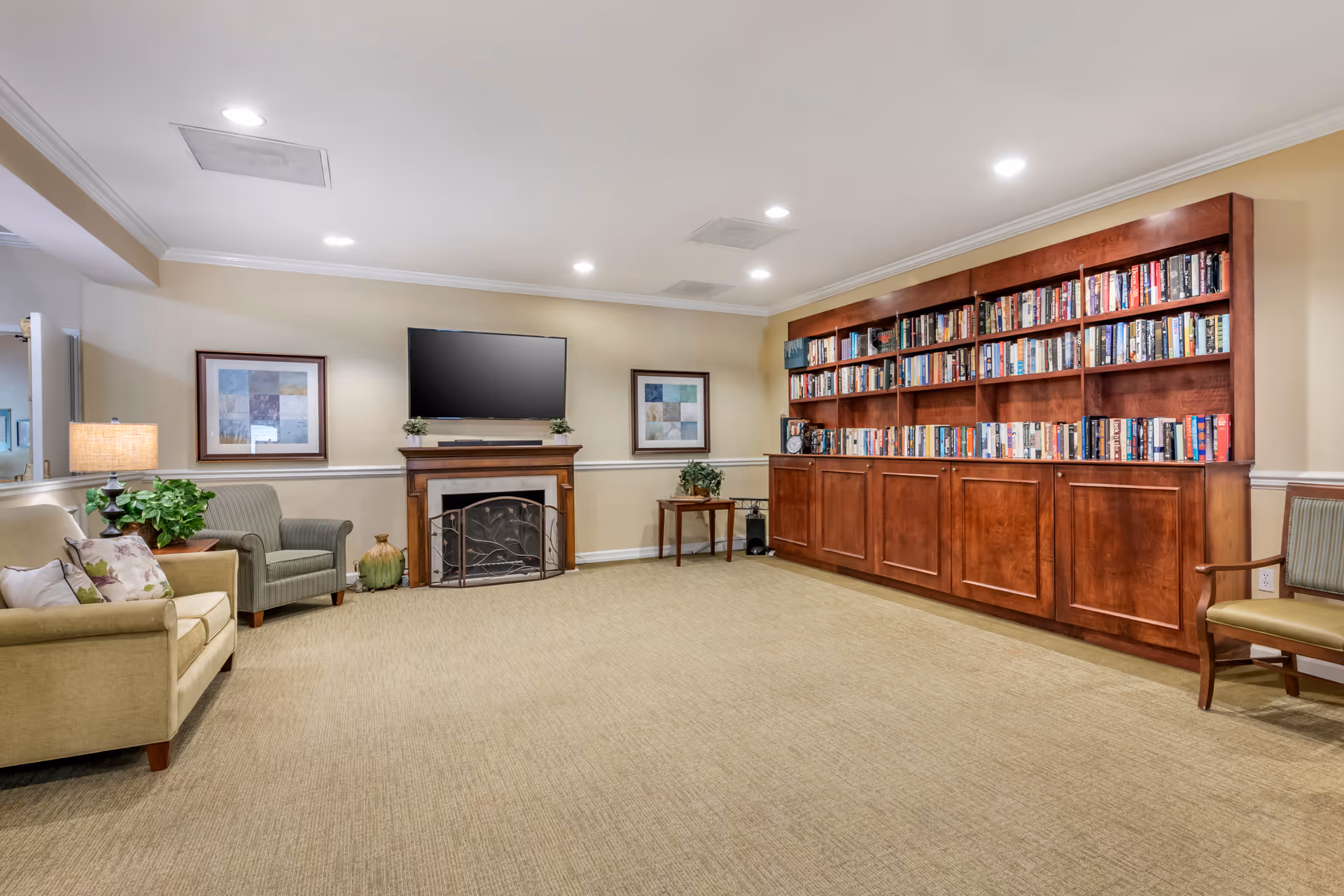 Cozy common room with sofas and chairs, a fireplace with a mounted TV, and a large built-in wooden bookshelf.