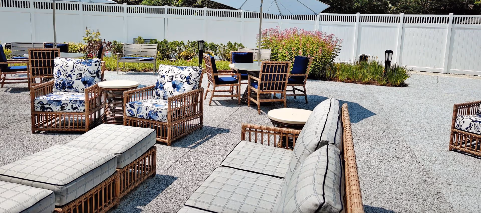 Outdoor patio area with wicker furniture including cushioned chairs and sofas, round tables with umbrellas, and a white privacy fence in the background. There are plants and flowers along the fence line.
