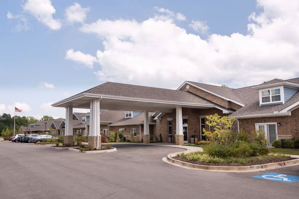 Exterior view of a senior living facility named Ciel of Collierville showing a covered entrance with brick walls, a driveway, parked cars, and an American flag in the background under a partly cloudy sky.