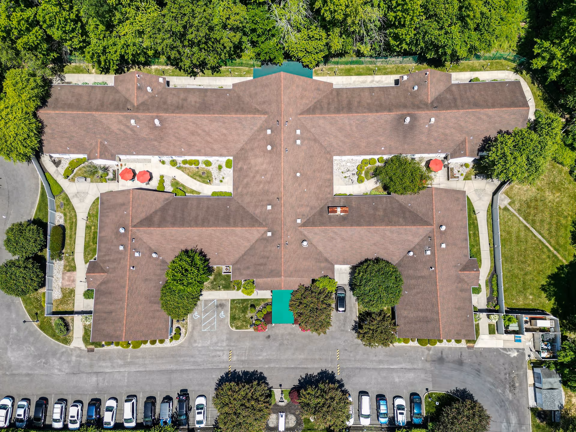 Aerial view of Arden Courts - ProMedica Memory Care Community (Cherry Hill) showing the building's roof, surrounding trees, parking lot with cars, and landscaped pathways with red umbrellas.