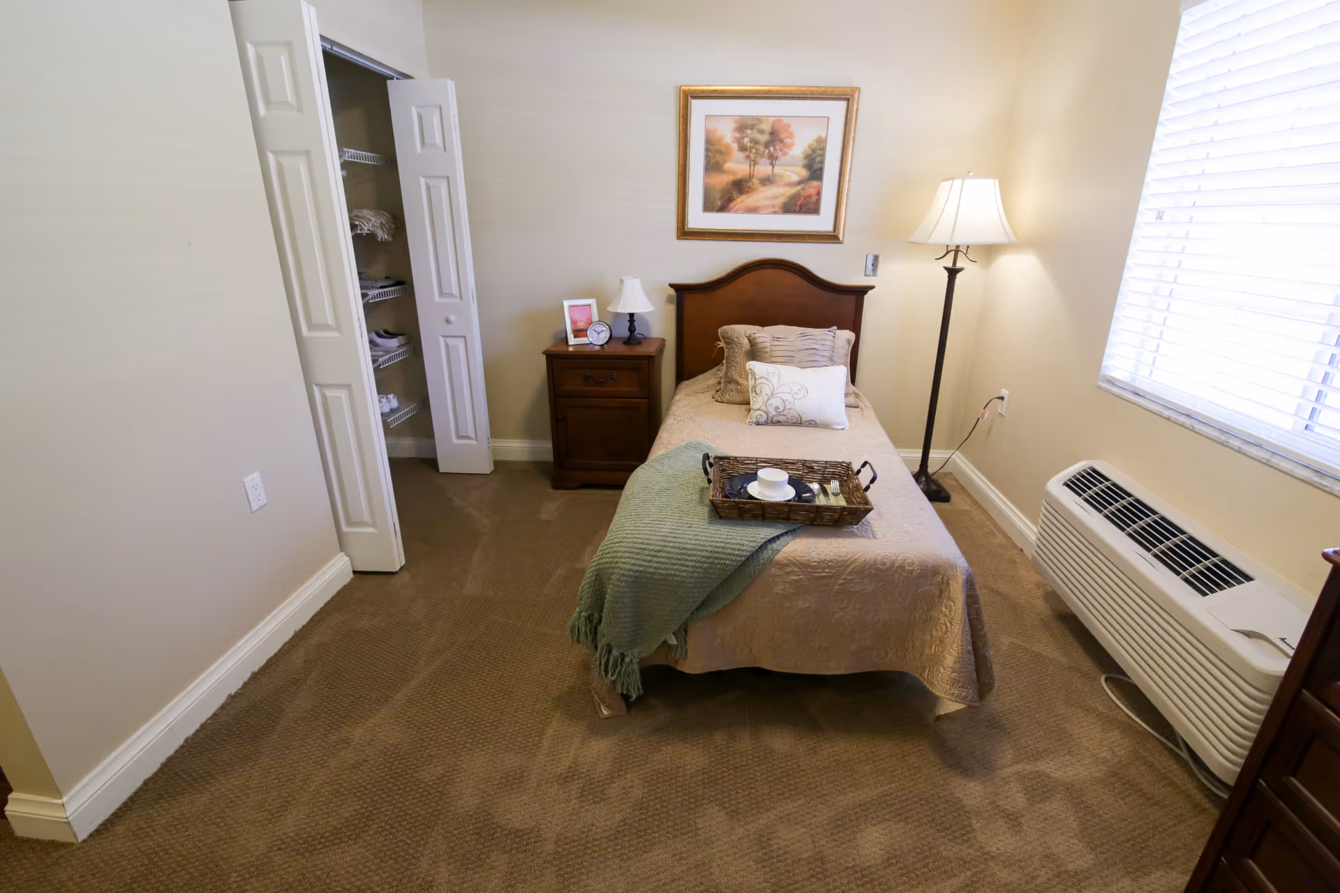 Small furnished bedroom with a single bed, nightstand, floor lamp, open closet, and a window air conditioner.