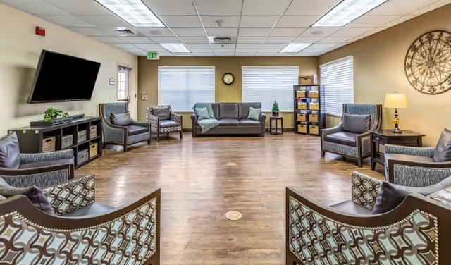 A spacious and well-lit living room with wooden flooring, multiple armchairs arranged in a circle, a sofa against the far wall, a wall-mounted flat screen TV, a small side table with a lamp, and decorative items including a large wall clock and a small plant. The room has large windows with blinds allowing natural light to enter.