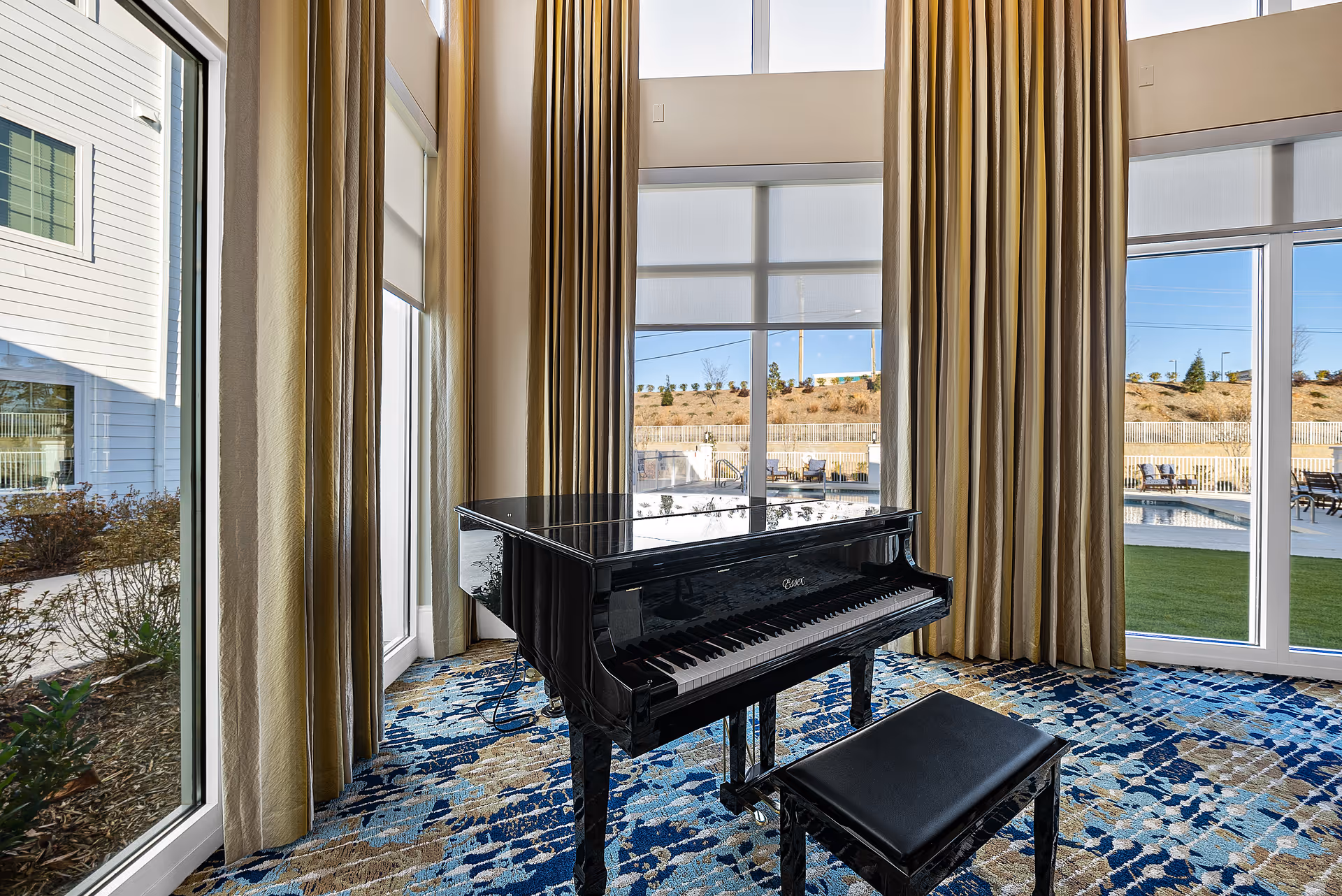 A black grand piano with a matching bench sits on a patterned blue and beige carpet in a room with tall windows and beige curtains. Outside the windows, there is a view of a pool area and a landscaped hill under a clear blue sky.