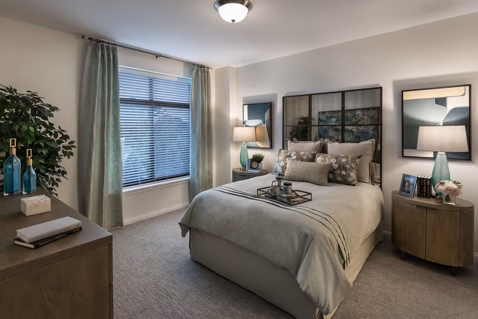 A cozy bedroom with a neatly made bed featuring multiple pillows and a decorative tray. The room has a large window with closed blinds and light green curtains. There are two bedside tables with matching lamps, a dresser with books and decorative items, and a potted plant in the corner. The walls are light-colored, and the room has a calm, inviting atmosphere.