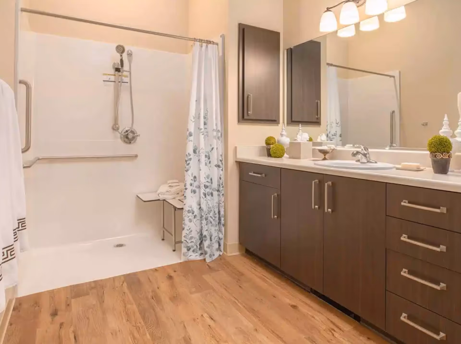 Accessible bathroom with a walk-in shower and floral curtain, wood-tone vanity with sink and mirror, and wood-look flooring.