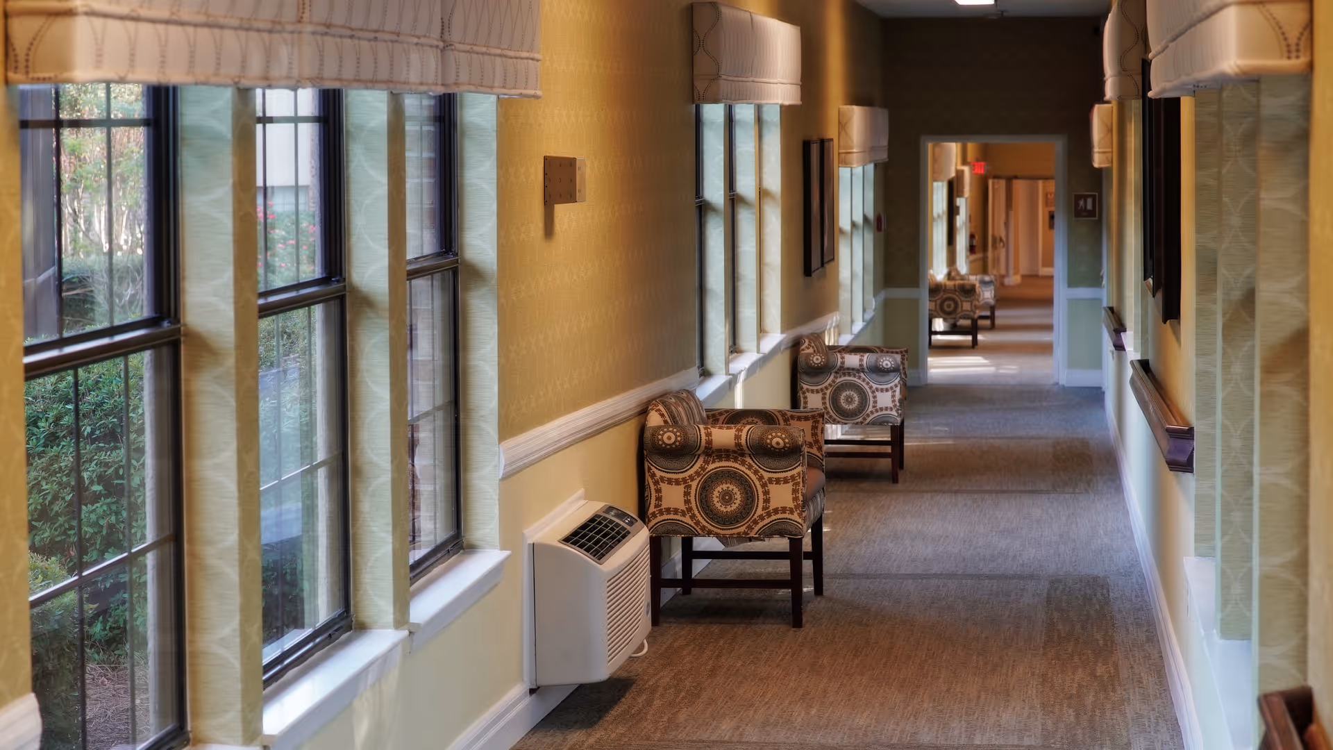 A long, carpeted hallway with large windows on the left side letting in natural light. Several patterned armchairs are placed along the wall beneath the windows. The walls are painted yellow with a white chair rail and have light-colored curtains above the windows. The hallway extends into the distance with more chairs and doorways visible.