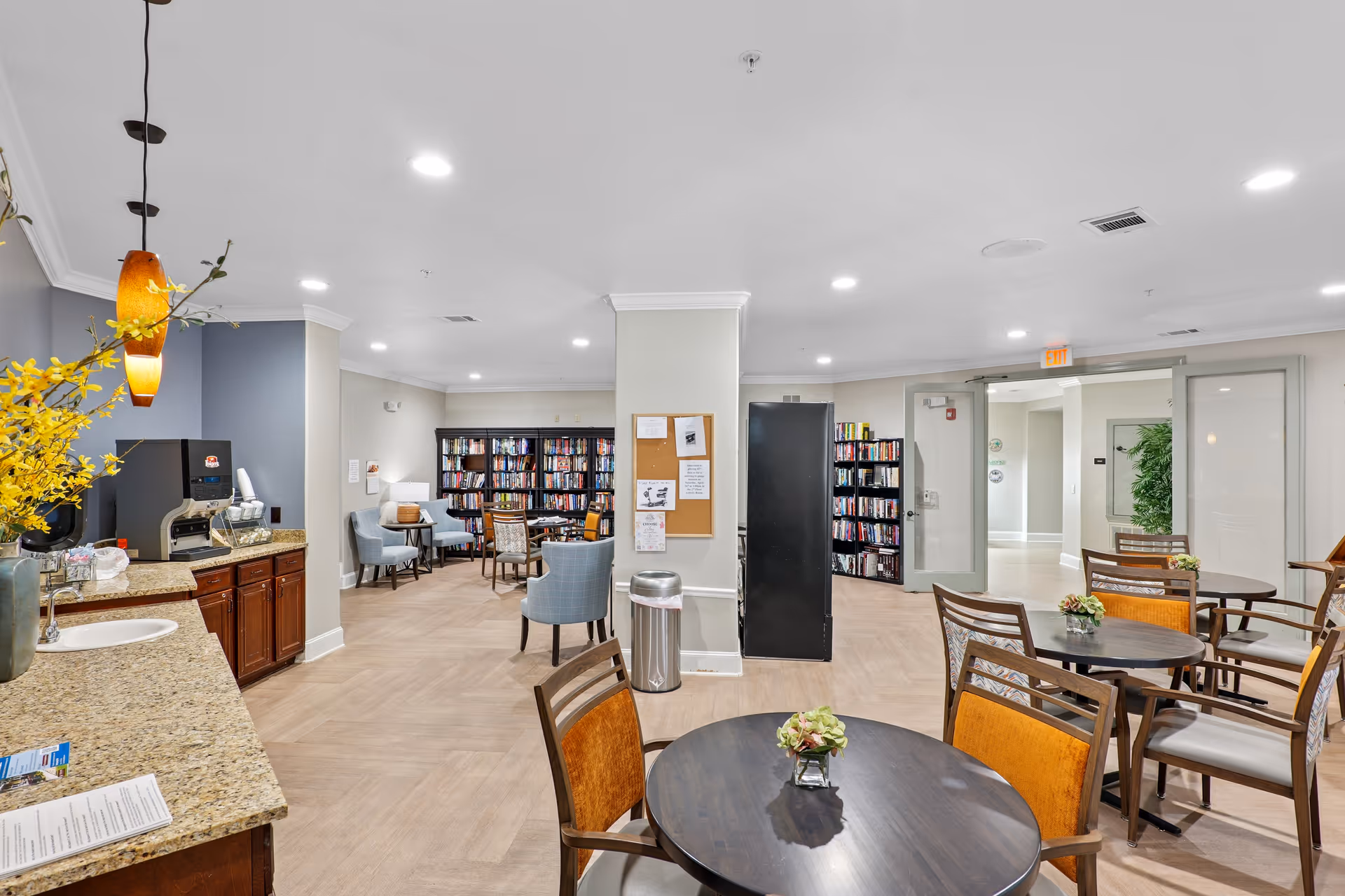 A bright and spacious common area in a senior living facility featuring a small kitchenette with granite countertops and a coffee machine on the left. Several round tables with chairs are arranged in the foreground and midground, each table decorated with a small flower arrangement. In the background, there is a cozy reading nook with bookshelves filled with books and comfortable armchairs. The room has light-colored walls, recessed ceiling lights, and a bulletin board on a central column. Glass double doors lead to another area of the facility.
