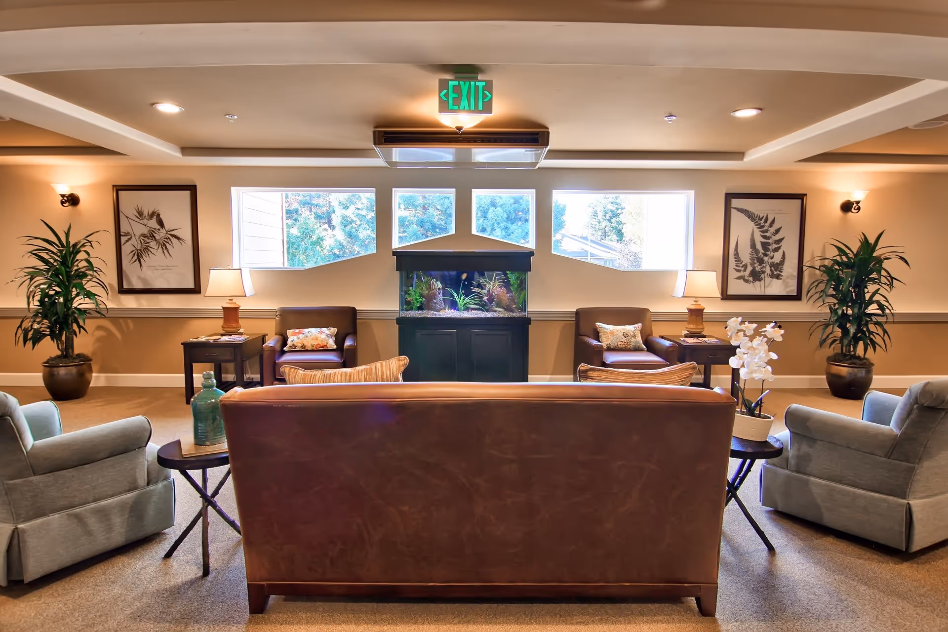 A cozy senior living common area with a brown leather sofa facing an aquarium. Two brown armchairs with floral cushions are placed on either side of the aquarium, each next to a small table with a lamp. Two gray armchairs flank the sofa, with a small round table holding a white orchid in front. The room has beige walls, carpeted floor, framed botanical prints, and potted plants. Three windows behind the aquarium let in natural light, and an illuminated green exit sign is above the aquarium.