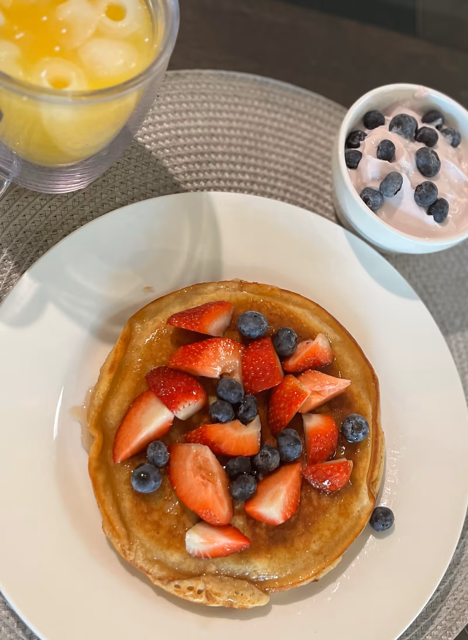 A plate with a pancake topped with fresh strawberries and blueberries, accompanied by a small bowl of pink yogurt with blueberries and a glass of orange juice with ice, all placed on a woven placemat.