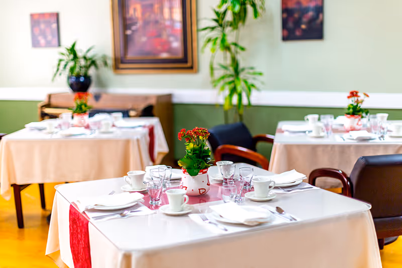 A dining room in a senior living facility with tables covered in white and beige tablecloths, each set with cups, glasses, napkins, and silverware. Small potted plants with red flowers serve as centerpieces. Chairs with wooden armrests surround the tables, and there are framed pictures and green plants on the walls.