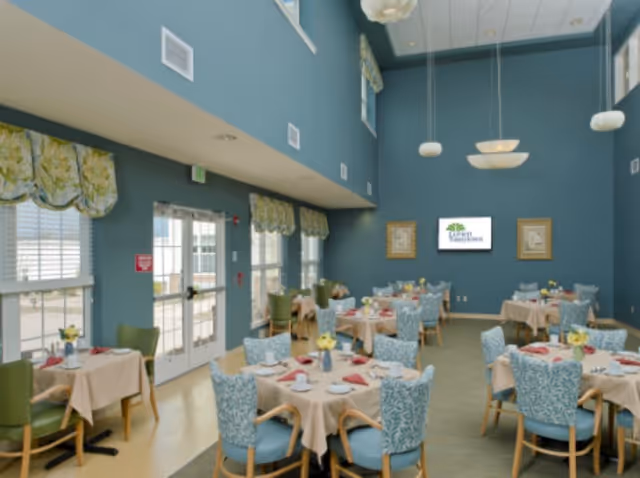 A spacious dining room with high ceilings painted in blue. Several round tables are set with beige tablecloths, white plates, cups, and red napkins. Blue patterned chairs and some green chairs surround the tables. Large windows with floral valances allow natural light to enter. A wall-mounted screen displays the Lorien Taneytown logo, flanked by two framed pictures. Modern pendant lights hang from the ceiling.