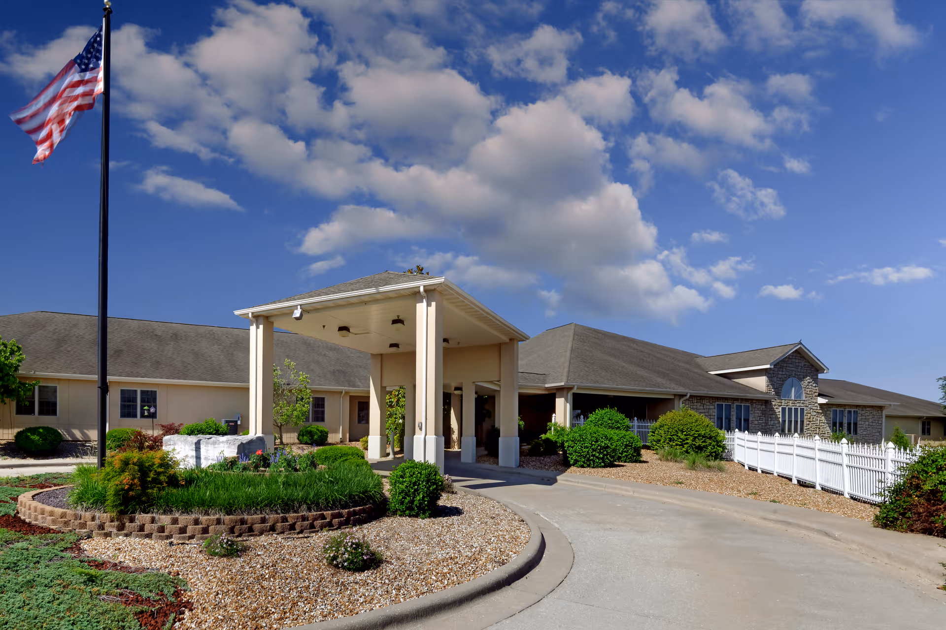 Exterior view of Ash Grove Healthcare Facility & Rehabilitation showing a single-story building with a covered entrance, landscaped garden with shrubs and flowers, an American flag on a flagpole, and a partly cloudy blue sky.