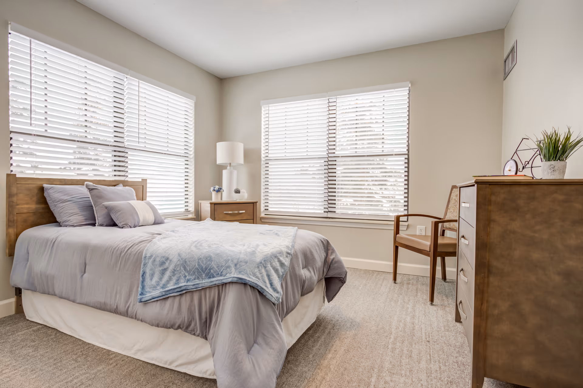 Bright furnished bedroom with a made bed, nightstand, dresser, chair, and large windows with blinds.
