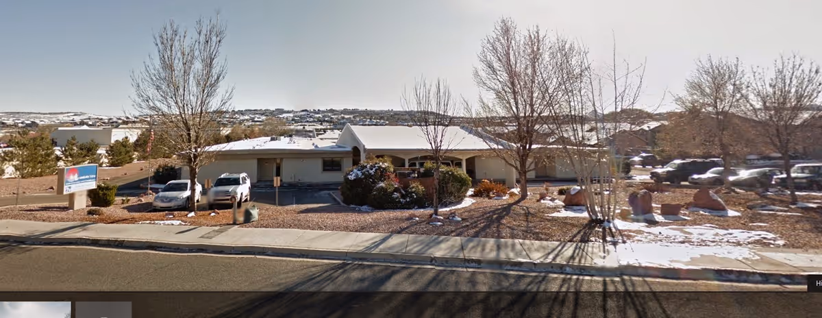 Exterior view of a single-story building with a covered entrance, surrounded by leafless trees and some snow on the ground. Several cars are parked near the building, and a sign is visible on the left side of the image. The sky is clear.