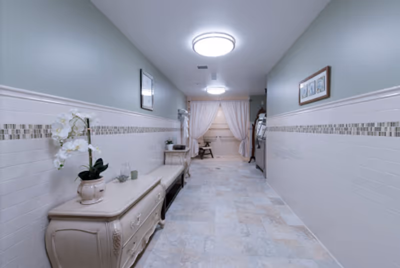 A long, narrow hallway with light-colored tiled floor and half-tiled walls with a decorative mosaic strip. On the left side, there is a cream-colored dresser with a white orchid plant and small decorative items. A bench is placed next to the dresser. The hallway is lit by two round ceiling lights and leads to a curtained area with a chair visible beyond the curtains. The walls are painted a soft grayish-green above the tiles, and framed artwork hangs on both sides.
