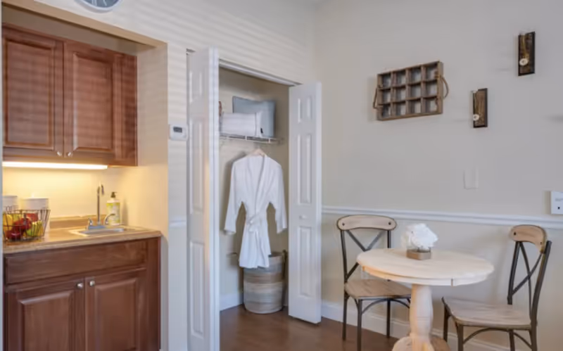 A small dining area with a round wooden table and two chairs with metal backs. To the left is a kitchenette with wooden cabinets, a sink, and a countertop. In the center background, there is an open closet with a white robe hanging inside and a basket on the floor. The walls are light-colored with simple decorative wall hangings.