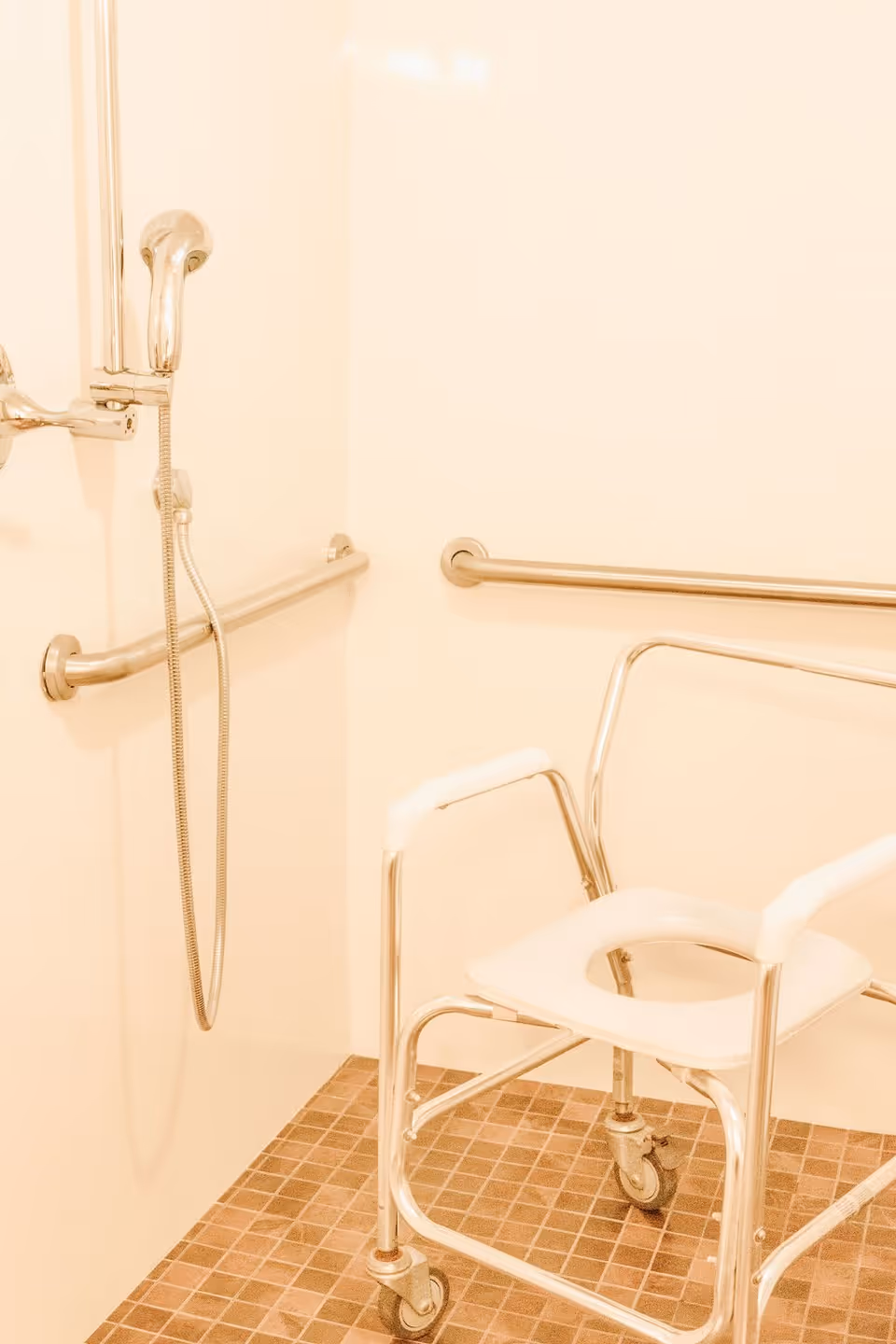 Accessible shower area with a handheld showerhead, two horizontal grab bars on the walls, and a mobile shower chair with armrests and wheels on a tiled floor.
