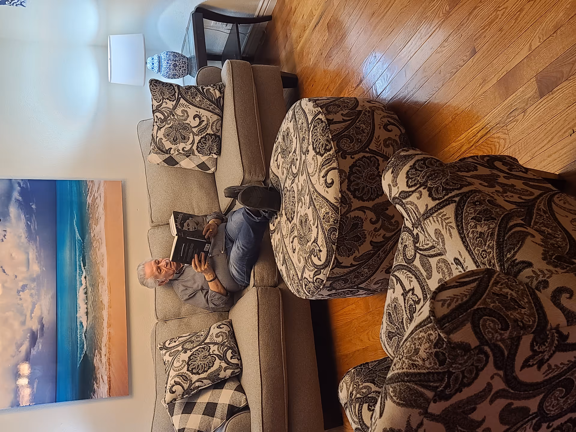An elderly man is sitting on a beige couch with patterned cushions, reading a book in a cozy living room. There is a matching patterned armchair and ottoman in front of him, a wooden floor, a side table with a blue and white lamp, and a large beach-themed painting on the wall behind the couch.