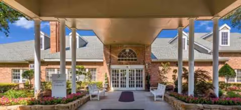 Brick senior living facility entrance with arched glass doors, columns, and flower beds.