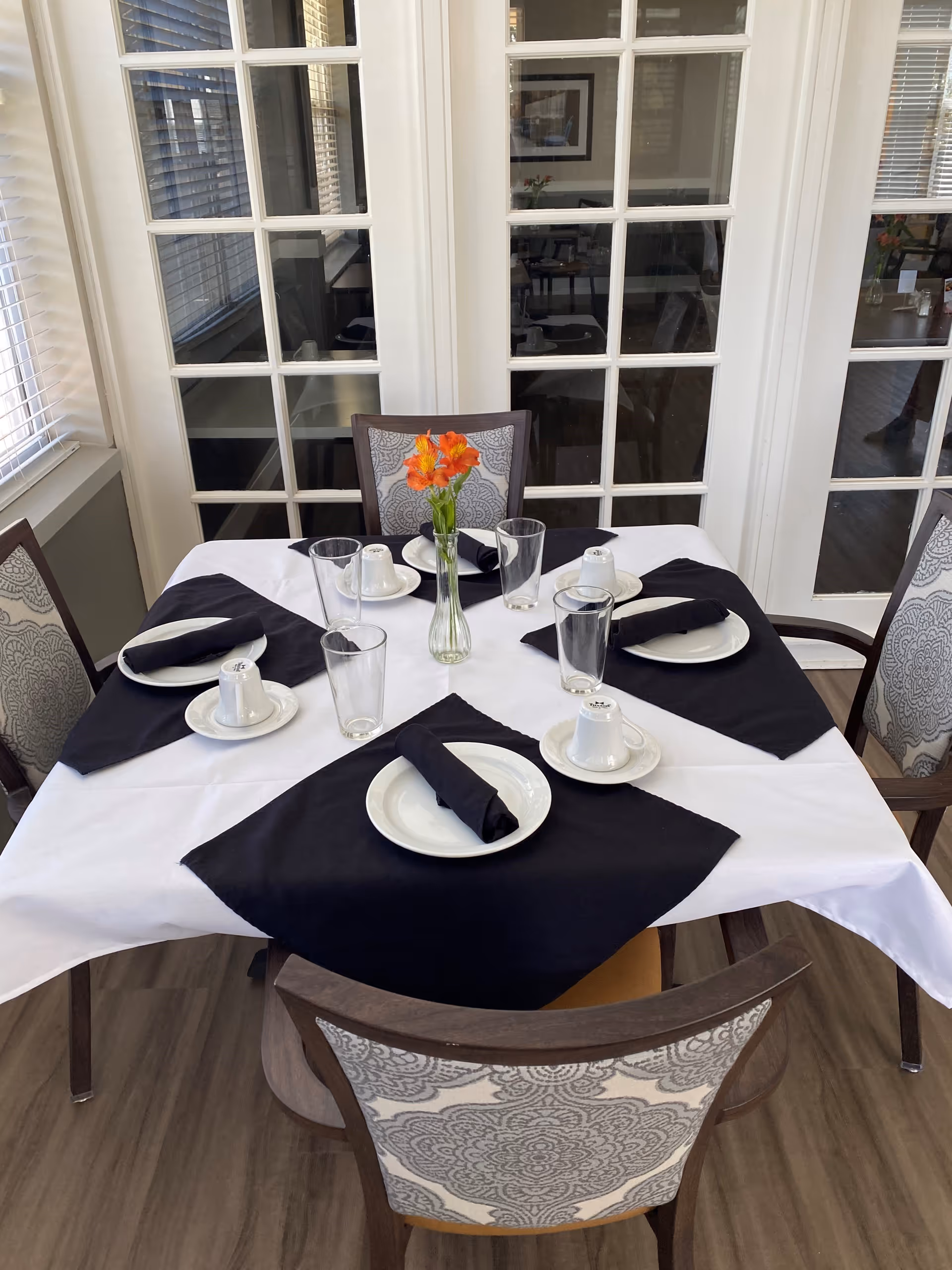A dining table set for four with white tablecloth and black napkins, each place setting includes a plate, a rolled black napkin, a glass, and an upside-down cup on a saucer. A small vase with orange flowers is placed in the center of the table. The table is surrounded by four chairs with patterned upholstery. Behind the table are white French doors with glass panes.