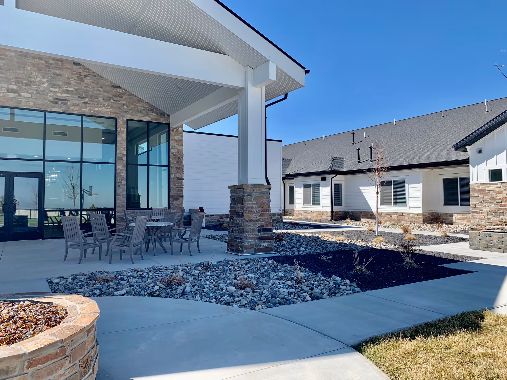 Covered outdoor courtyard with patio tables and chairs in front of a stone-accented assisted living building with large windows.
