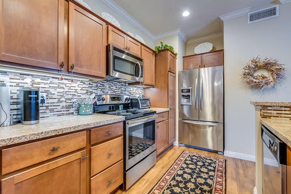 Modern kitchen with wooden cabinets, stainless steel appliances including a microwave, stove, and refrigerator. The backsplash features a mosaic tile design, and there is a patterned rug on the wooden floor. A decorative wreath hangs on the wall near the refrigerator.