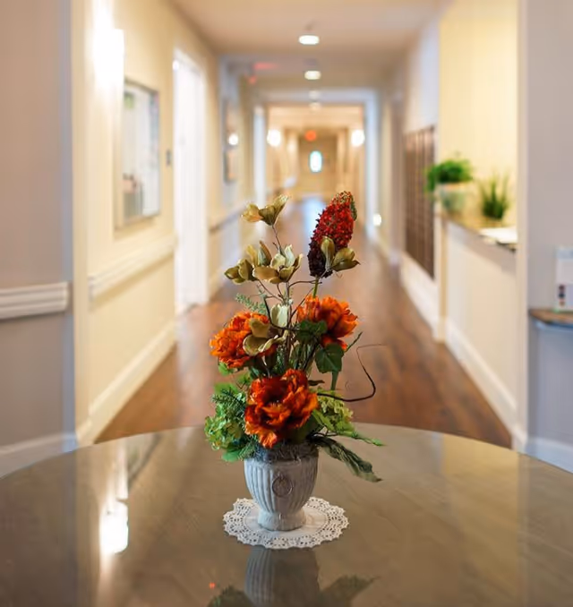 A decorative flower arrangement with orange and red flowers in a white vase placed on a round table with a lace doily, set in a well-lit hallway with wooden flooring and light-colored walls in a senior living facility.