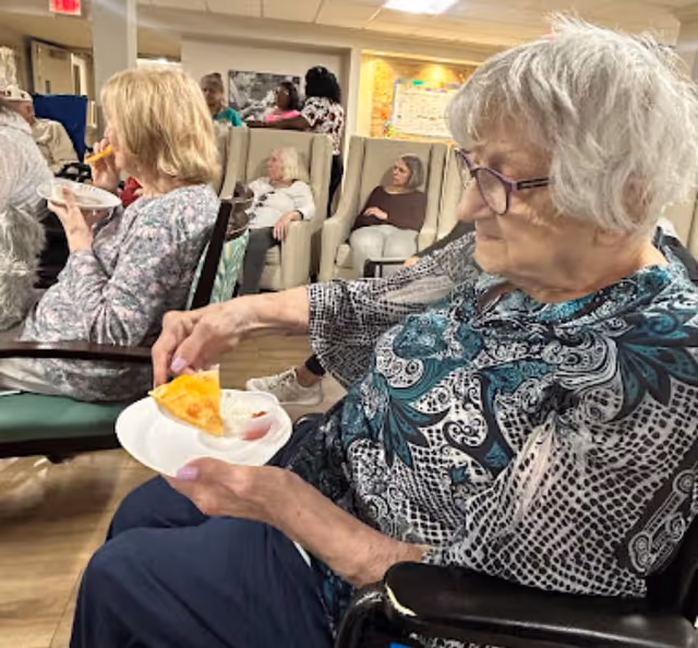 An elderly woman with gray hair and glasses sits in a wheelchair holding a plate with a slice of pizza and dipping sauce. Other elderly individuals are seated in the background in a communal room with chairs and a bulletin board on the wall.