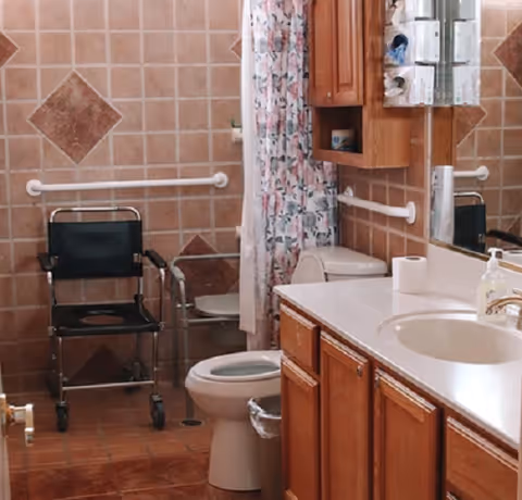 Bathroom with brown tiled walls and floor, featuring a toilet, a shower area with a floral shower curtain, a black shower chair, grab bars on the walls, a wooden cabinet above the toilet, and a sink with a countertop and mirror.