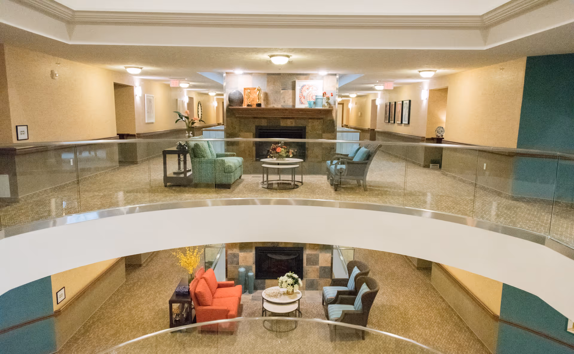 Interior view of a senior living facility lounge area with two levels. The upper level has two green armchairs and a small round table with flowers in front of a fireplace. The lower level has a red sofa, two blue armchairs, and a round table with flowers in front of another fireplace. The space is well-lit with ceiling lights and decorated with artwork and plants.