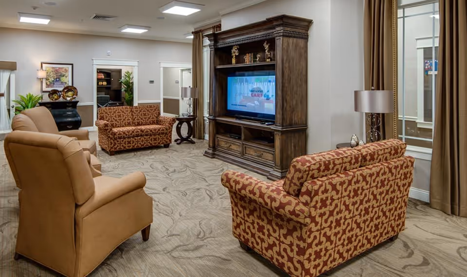 A cozy senior living facility common area with patterned sofas and beige armchairs arranged around a large wooden entertainment center with a flat-screen TV. The room has beige walls, carpeted flooring, framed artwork, potted plants, and large windows with brown curtains.