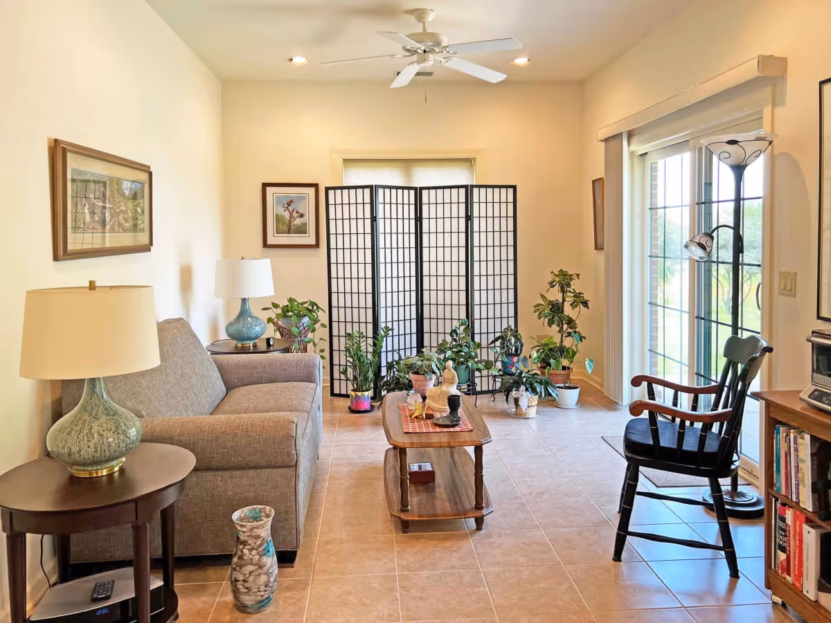 A cozy living room with a gray sofa, two side tables with lamps, a wooden coffee table with decorative items, several potted plants near a folding screen, a black wooden chair, a floor lamp, and a sliding glass door leading outside.