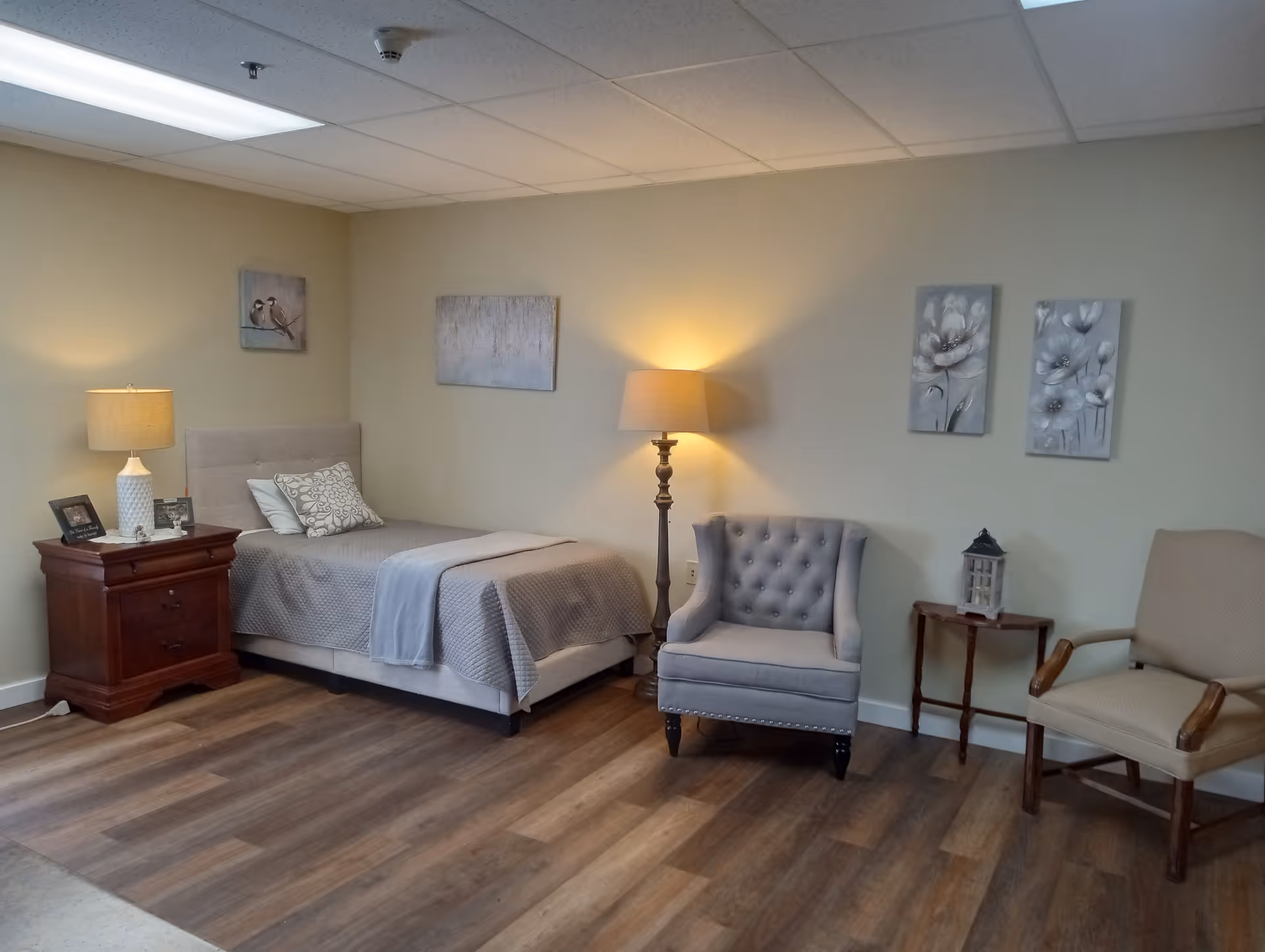 Furnished bedroom with a single bed, wooden nightstand, lamps, and two chairs against a wall with artwork.
