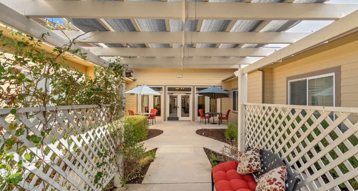 Outdoor patio area with a pergola overhead, lattice fencing on both sides, and a concrete walkway leading to a building entrance. There are tables with umbrellas and red chairs, along with a cushioned bench with patterned pillows on the right side.