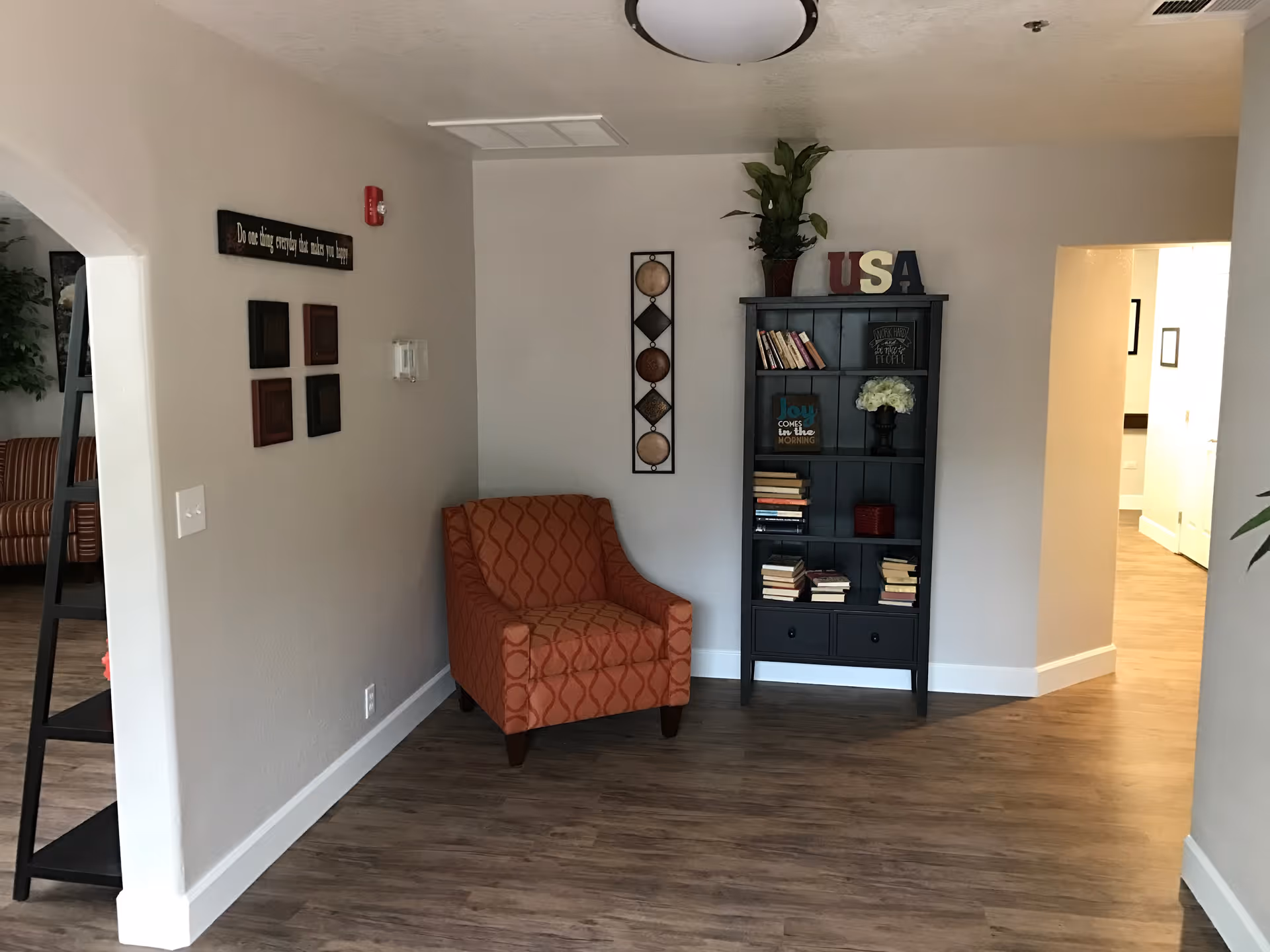 A cozy corner of an assisted living facility featuring a patterned orange armchair next to a black bookshelf filled with books and decorative items, including a plant and letters spelling USA. The room has light-colored walls, wood flooring, and a ceiling light fixture. There is an open doorway leading to another room with more light and furniture visible.