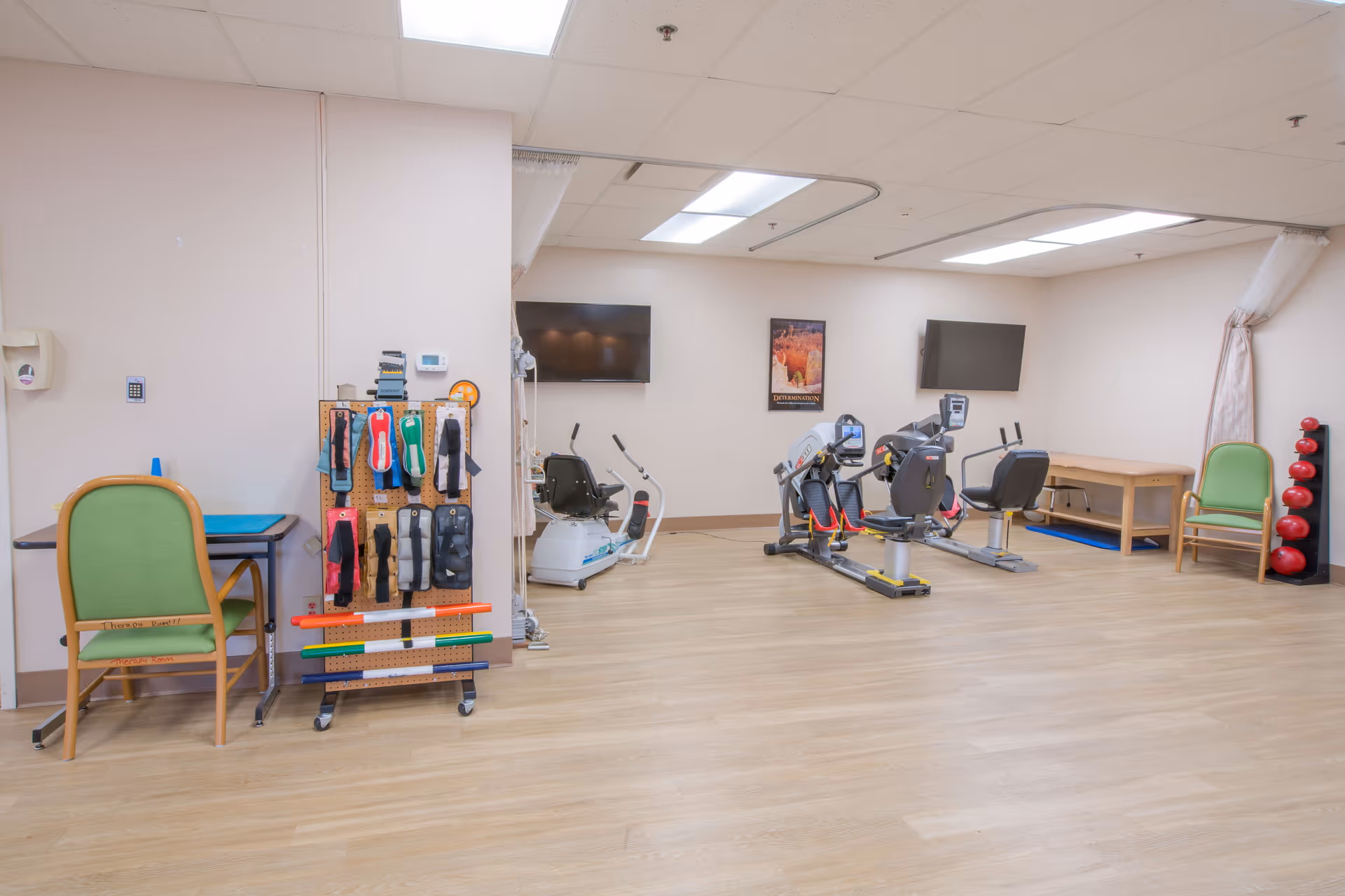 A spacious rehabilitation or physical therapy room with exercise equipment including stationary bikes and weights. The room has light wood flooring, white walls, and ceiling lights. There are two green chairs, a small table, and a rack holding various therapy tools and exercise bands. Two flat-screen TVs are mounted on the walls.