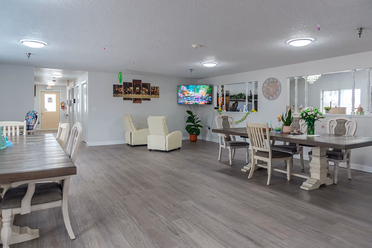 A spacious, well-lit common area in an assisted living facility featuring two wooden dining tables with chairs, two cream-colored recliners facing a wall-mounted TV, a large wall clock, and decorative plants and flowers. The room has light gray walls and wood-look flooring.