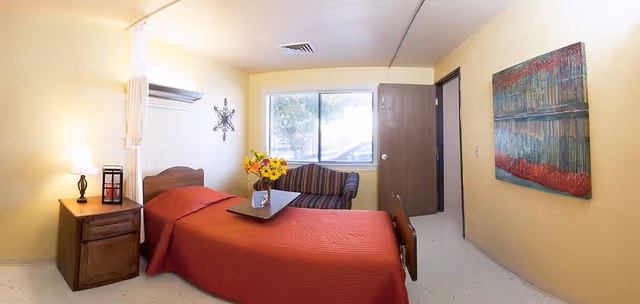 A small, cozy bedroom with a single bed covered in a red bedspread. Next to the bed is a wooden nightstand with a lamp and a decorative lantern. A vase with yellow flowers sits on a tray on the bed. There is a window with a view of trees, a striped armchair beneath the window, and a colorful abstract painting on the wall. The room has light yellow walls and a door leading to another area.