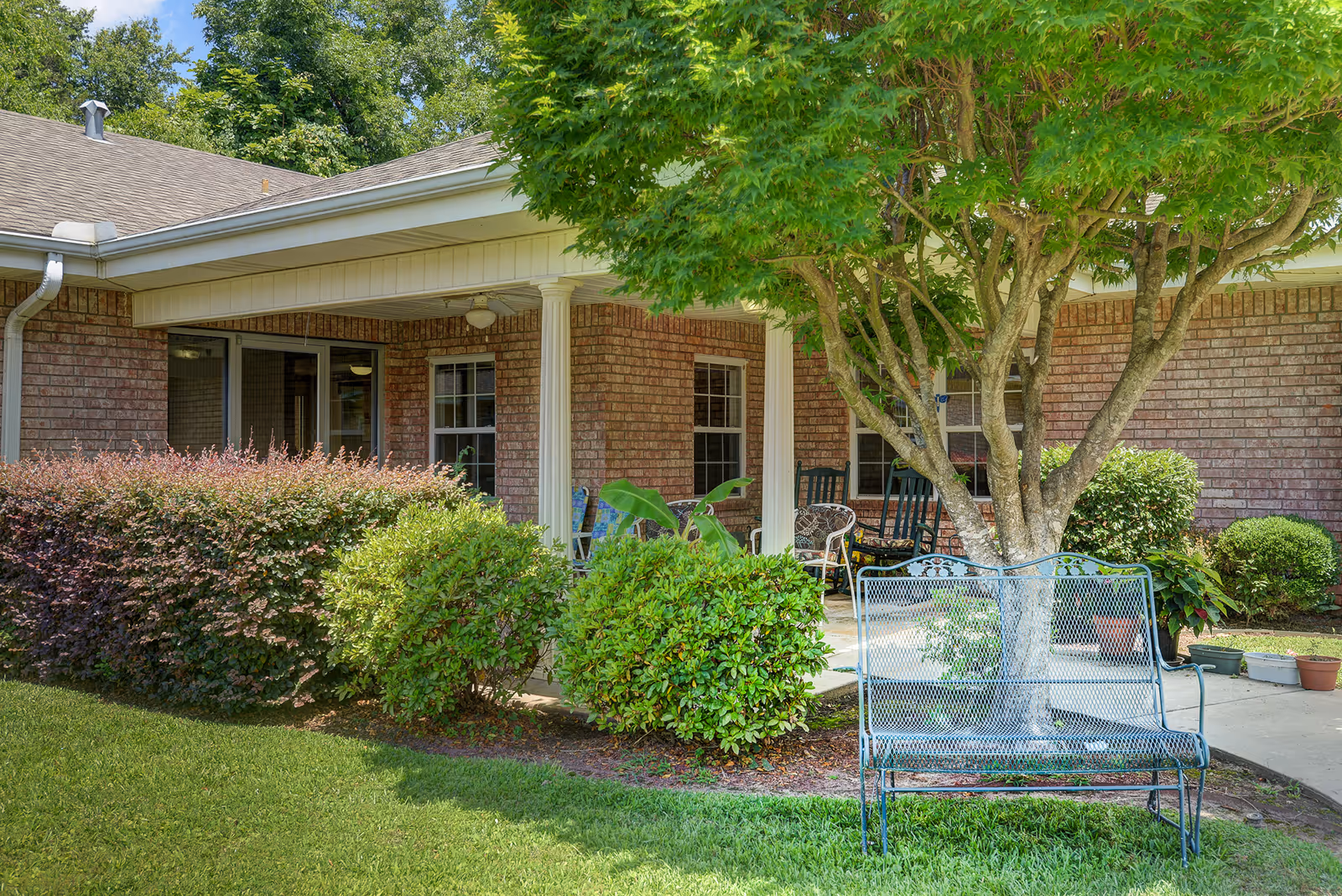 Outdoor patio area of Oakwood House with a brick exterior, white columns, green bushes, a tree, a metal bench, and several chairs on the covered porch.