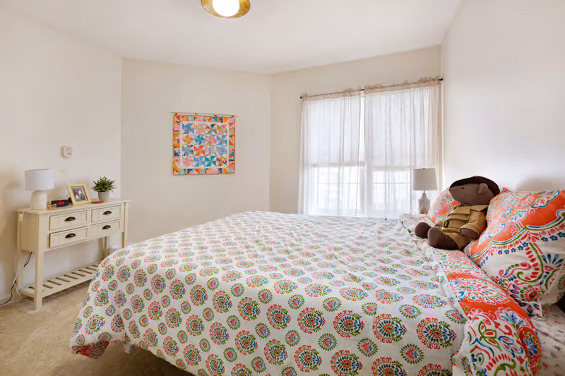 A bright bedroom with a large bed covered in a colorful patterned bedspread and matching pillows. A stuffed bear sits on the bed near the pillows. There is a window with sheer curtains allowing natural light to fill the room. On the left side, there is a small white table with drawers, a lamp, a framed photo, and a small potted plant. A colorful quilt hangs on the wall above the table.