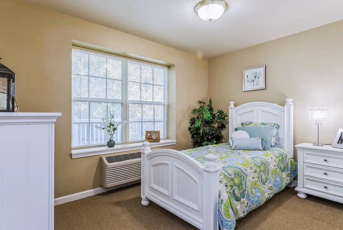 A bedroom with a white wooden bed frame and a colorful green and blue paisley patterned bedspread. There are multiple pillows on the bed. Next to the bed is a white nightstand with a lamp and a framed photo. A large window with blinds lets in natural light. A green potted plant is in the corner of the room, and a framed floral picture hangs on the beige wall above the bed.