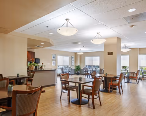 Spacious senior dining room with multiple round tables and chairs, pendant lighting, wood floors, and large windows.