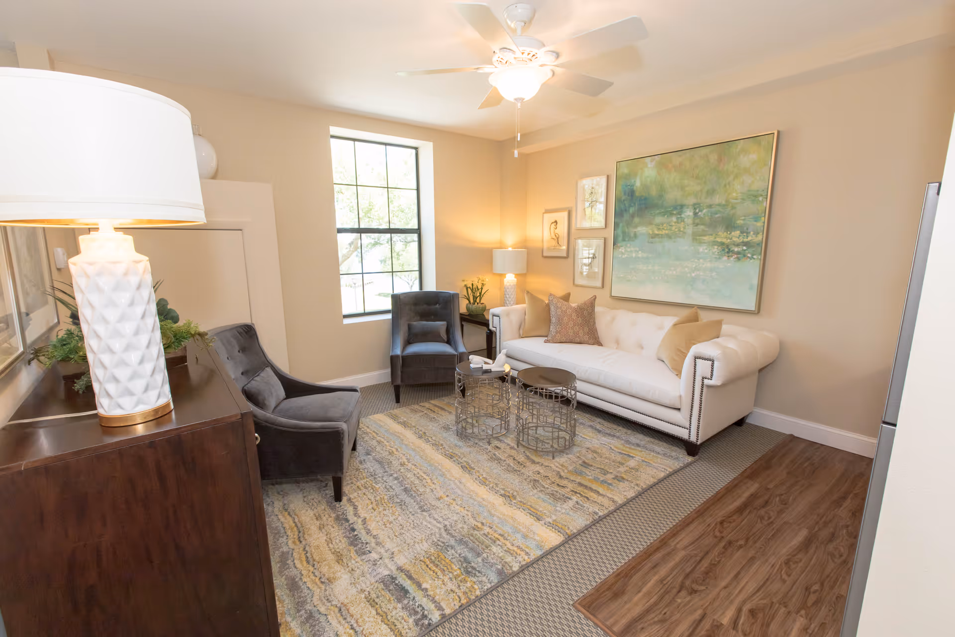 A cozy living room with a white tufted sofa adorned with beige and patterned pillows, two dark gray armchairs, a patterned area rug, two round metal coffee tables, a large window letting in natural light, a ceiling fan with light, and various framed artworks on the walls. A wooden cabinet with a white textured lamp and green plants is also visible.