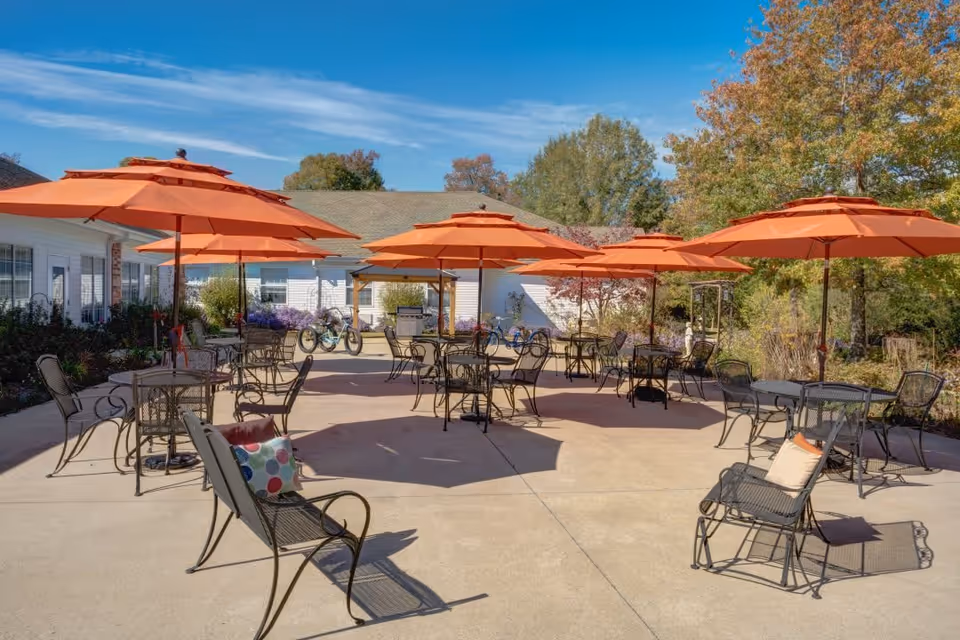 Outdoor patio area with multiple black metal tables and chairs, each shaded by large orange umbrellas. The patio is surrounded by greenery and trees with autumn foliage, and a white building is visible in the background under a clear blue sky.