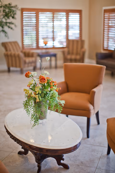 A cozy living room area with a white marble oval coffee table holding a vase of orange and yellow flowers with green foliage. Surrounding the table are several upholstered chairs in warm tones. The background features large windows with wooden blinds and additional seating.